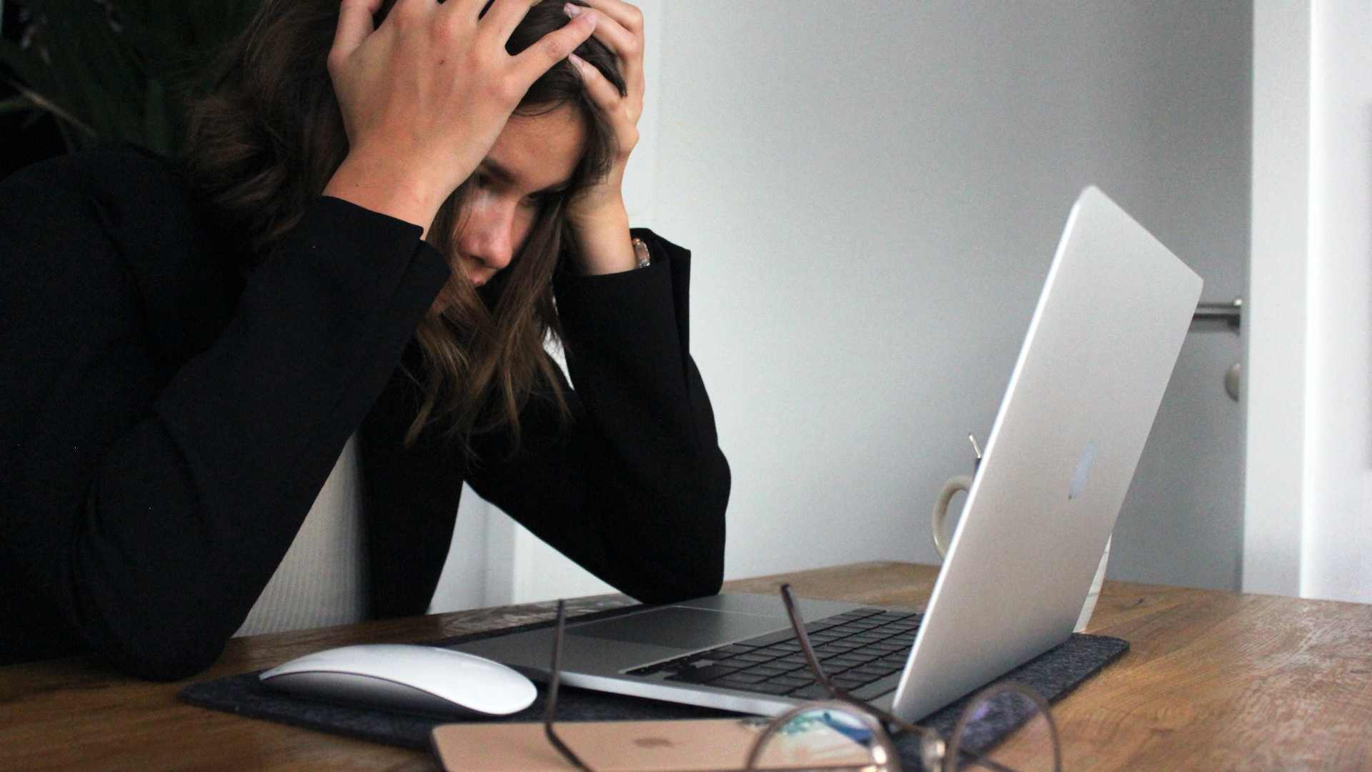 A woman sitting at a desk with a laptop, with her hands on her head looking stressed 